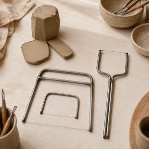 May include: A set of three metal wire clay cutters with silver-toned frames and thin cutting wires. The collection includes two rectangular-shaped wire tools and one T-shaped wire tool, all designed for slicing through blocks of raw, light brown clay.