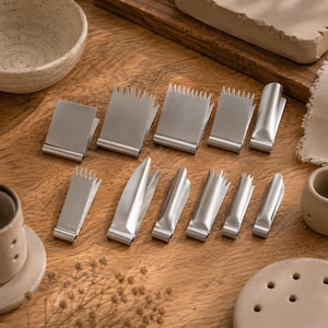 May include: A collection of ten silver-coloured metal clips arranged in two rows. The top row features four clips with serrated edges and one smooth clip. The bottom row contains five clips of varying widths, including serrated and smooth-edged designs.