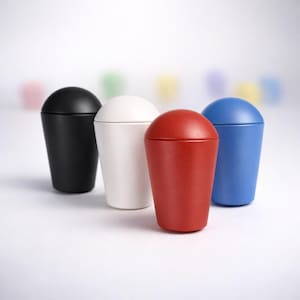 May include: Four lidded containers in black, white, red, and blue. The containers have a rounded top and a tapered body. The background features blurred, pastel-coloured shapes. The containers are likely for storage or decorative purposes.