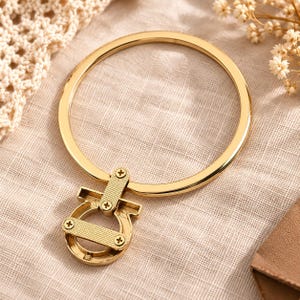 May include: A gold-toned metal circular key ring featuring a decorative Gancini-style clasp attachment. The clasp is secured with four small screws and has a textured, cross-hatched finish on the horizontal bar, while the ring itself is smooth.