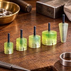 May include: A set of five translucent yellow ring stretchers with black metal mandrels. Four of the tools are cylindrical with stepped, grooved sides of varying diameters, and one is a tapered, ribbed cone shape for shaping metal rings.