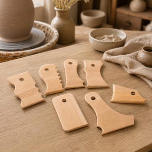 May include: A set of seven light-colored wooden pottery ribs and scrapers, each featuring a small circular hole for hanging. These tools exhibit various shapes, including serrated edges, curved profiles, and straight sides, designed for shaping clay.