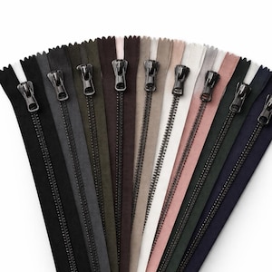May include: A selection of coloured zippers, including black, grey, olive green, brown, beige, white, and pink. Each zipper has a metal pull and is displayed on a matching fabric strip. Suitable for sewing and craft projects.