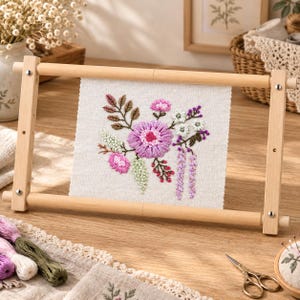 Peut inclure: Un carré de tissu clair présentant une broderie florale avec des fils roses, violets et verts, maintenu dans un cadre rectangulaire en bois clair. La broderie représente une fleur centrale entourée de petites fleurs et de feuilles.