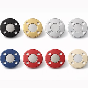 Pode incluir: Uma coleção de oito fechos magnéticos redondos em várias cores. Os fechos têm um disco central prateado rodeado por um anel colorido com seis orifícios espaçados uniformemente. As cores incluem preto, dourado, prateado, azul, vermelho e bege.