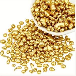 May include: A collection of small, irregular, metallic gold-colored nuggets scattered across a surface, with a larger cluster contained within a white bowl. The pieces have a smooth, reflective, and polished surface with varying shapes and sizes.