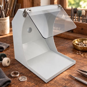 May include: A white metal dust collection hood with a clear acrylic shield attached by two metal hinges. The device features a curved back panel, a flat base, and a circular opening on the interior wall, designed for jewelry polishing and grinding tasks.