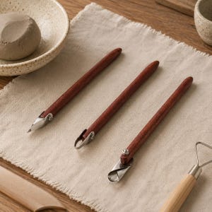 May include: Three pottery carving tools with dark reddish-brown wooden handles and silver-coloured metal tips. Each tool features a distinct metal blade shape, including a pointed tip, a small loop, and a curved scoop, all secured with metal rivets.