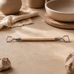 May include: A double-ended pottery ribbon tool featuring a smooth, light-coloured wooden handle and two silver-toned metal wire loops at each end, designed for carving and shaping clay with precision during the ceramic sculpting process.