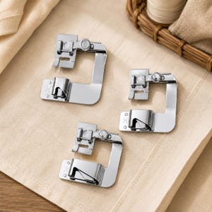 Pode incluir: Três calcadores de bainha para máquina de costura em metal prateado, cada um com uma estrutura em forma de U, um pequeno parafuso de ajuste e uma guia para bainha enrolada. Os componentes metálicos têm um acabamento polido.