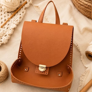 Puede incluir: Una mochila de cuero color canela con una solapa redondeada, un asa superior y un cierre de presión de metal dorado. Los bordes de la bolsa están perforados con pequeños agujeros para coser y la estructura se mantiene con remaches dorados.