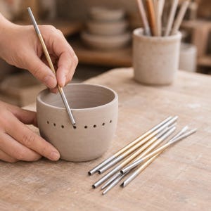 Peut inclure: Un bol en céramique beige clair avec une rangée de petites perforations circulaires près du bord, accompagné de plusieurs outils métalliques fins dotés d'une pointe argentée et d'un manche doré, utilisés pour le travail de l'argile.