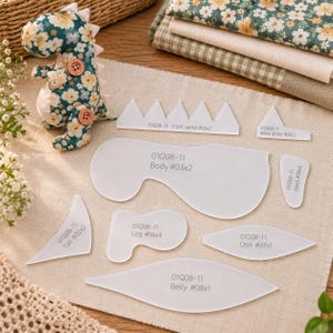 May include: A stuffed dinosaur toy with a dark green floral pattern and two brown buttons, accompanied by a set of clear acrylic sewing templates labelled with text including 01Q08-11, Body #03x2, Tail #02x2, Leg #06x4, Chin #07x1, Belly #08x1, Hand #08x4, and spine pieces.