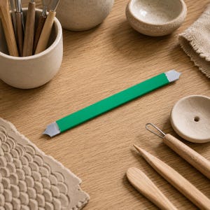 Puede incluir: Una herramienta de alfarería de doble extremo con un mango rectangular de color verde vibrante y dos puntas de metal plateado con extremos puntiagudos y muescas. La herramienta se sitúa en el centro entre varios utensilios de madera y cuencos de cerámica.