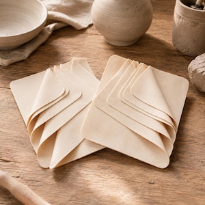 May include: Two stacks of light beige, rectangular, thin, flexible sheets arranged in a fan-like pattern. Each stack consists of multiple layers of smooth, matte material with rounded corners, overlapping to create a layered, folded appearance.
