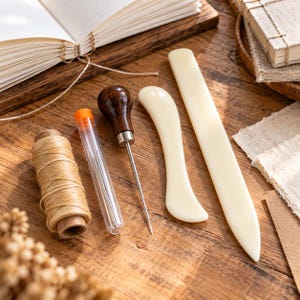 Könnte beinhalten: Ein Buchbindewerkzeug-Set bestehend aus einer Spule mit beigem Garn, einem durchsichtigen Kunststoffröhrchen mit Nadeln, einer Ahle mit Holzgriff, einem gebogenen Falzbein und einem langen, geraden Falzbein für Papierarbeiten.