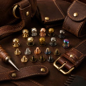 May include: A selection of metal rivets in gold, silver, copper, and iridescent finishes, arranged on a brown leather surface. The rivets feature a domed head and a threaded post. Leather accessories, such as a bag, wallet, and belt, are also visible.