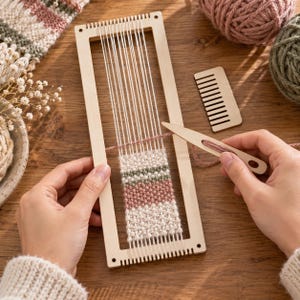 Op de afbeelding: Een rechthoekig houten weefraam met witte scheringdraden en een gedeeltelijk geweven textiel met horizontale strepen in crème, oudroze en saliegroen. Een houten weefspoel en een kleine houten kam zijn inbegrepen.