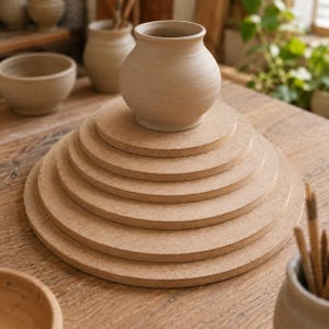 Op de afbeelding: Een stapel van zes ronde, lichtbruine houten planken gerangschikt in afnemende grootte, bekroond met een kleine, ongeglazuurde beige keramische vaas. De planken hebben een glad, vlak oppervlak en een consistente, natuurlijke houtnerftextuur.
