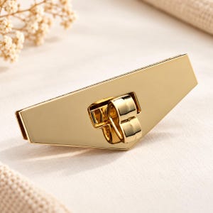 Könnte beinhalten: Ein goldfarbenes Taschenschloss aus Metall mit einer breiten, winkligen, trapezförmigen Platte und einem zentralen, polierten, U-förmigen Drehverschluss. Das Beschlagteil hat eine glatte, reflektierende Oberfläche und eine geometrische Form.