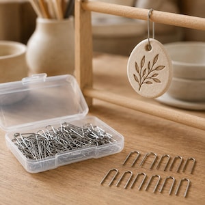 Pode incluir: Um ornamento oval de argila bege com um design de folha em relevo pende de um suporte de madeira por um gancho de metal prateado. Uma caixa de armazenamento de plástico transparente cheia de pinos de metal prateado em forma de U está ao lado.