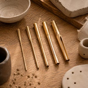 May include: Five cylindrical brass tools of varying diameters arranged in a row. Each tool features a smooth, polished metallic finish with a tapered end, and the largest tool includes a vertical slit along its upper section for specialised use.