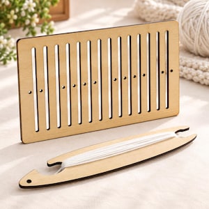 Peut inclure: Un métier à tisser rectangulaire en bois clair avec des fentes verticales et de petits trous circulaires, accompagné d'une navette en bois incurvée enroulée de fil blanc. Le métier présente une finition lisse et un design plat et structuré.