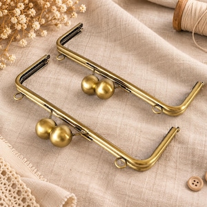 May include: Two rectangular antique brass purse frames with rounded corners. Each frame features a kiss-lock closure with two smooth, spherical brass beads and small metal loops attached to the sides for connecting straps or accessories.
