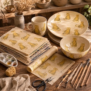 Op de afbeelding: Een set van een keramische mok, kom en bord met een geel en zwart luipaardpatroon, vergezeld door een stapel bijpassende vellen papier met luipaardprint, een verfpalet, een spons, een schaar en diverse houten boetseerwerktuigen op een tafel.