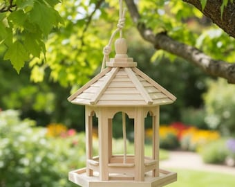 May include: A wooden bird feeder with a pagoda-style roof hangs from a tree branch. The natural wood feeder has an open design with arched openings and a tray base. A rope is used to suspend the feeder, which is surrounded by green foliage.
