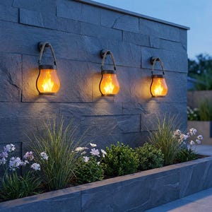 Puede incluir: Tres faroles de pared para exteriores con efecto llama. Cada farol tiene un marco de metal oscuro, un asa de cuerda y un brillo naranja cálido. Los faroles están montados en una pared de piedra gris oscuro sobre un lecho de vegetación y flores.