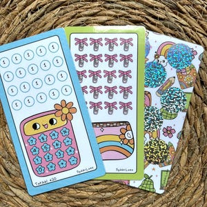 Op de afbeelding: Drie kleurrijke sticker vellen met leuke illustraties. Eén vel toont een rekenmachine met bloemaccenten en de tekst "Totaal €20". Een ander heeft roze strikken en de derde heeft regenbogen en glinsterende vormen. De stickers liggen op een geweven achtergrond.