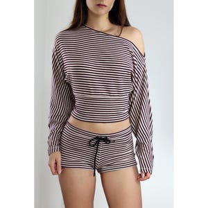 May include: A two-piece striped outfit. The long-sleeved crop top has an off-the-shoulder design, and the matching shorts have a drawstring. Both the top and shorts feature horizontal stripes in a dark color and white.