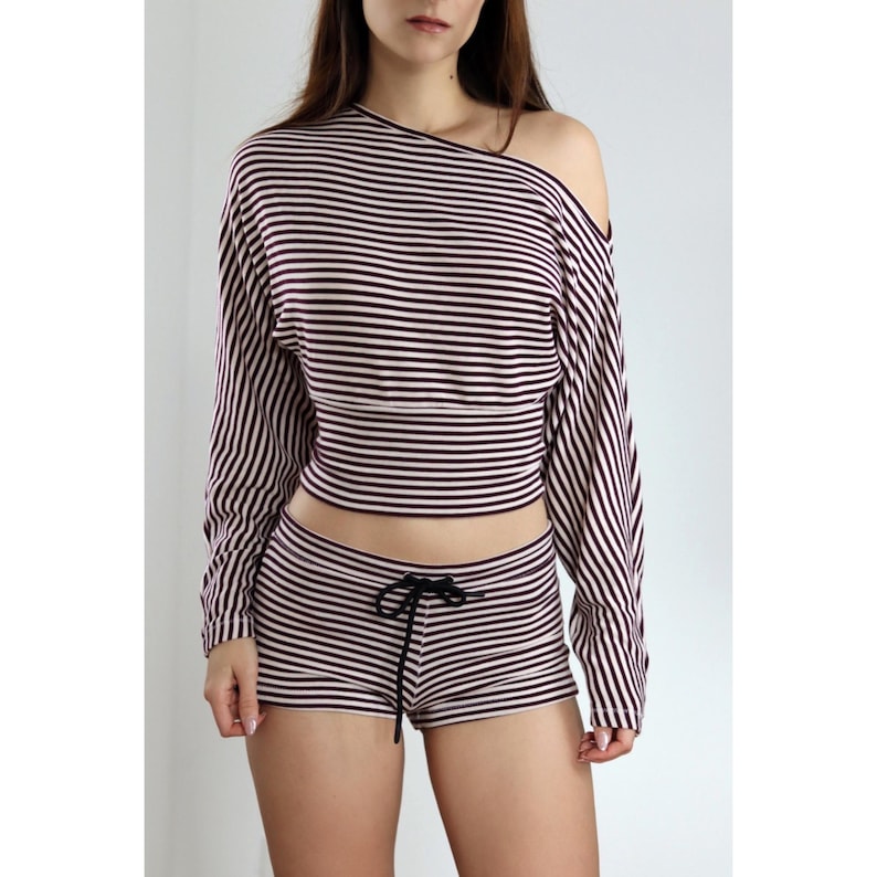 May include: A two-piece striped outfit. The long-sleeved crop top has an off-the-shoulder design, and the matching shorts have a drawstring. Both the top and shorts feature horizontal stripes in a dark color and white.