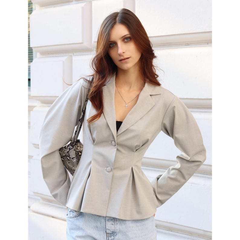 Puede incluir: Una blazer gris claro con cintura entallada y detalles plisados. La blazer tiene mangas largas y dos botones. La persona lleva un top negro y vaqueros azul claro. Se ve un peque&ntilde;o collar con una cruz.