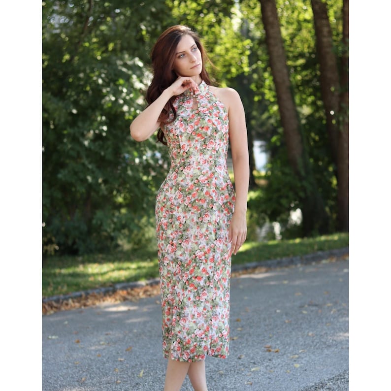 K&ouml;nnte beinhalten: Ein Halfterkleid mit Blumendruck und hohem Ausschnitt. Das Kleid hat ein Muster aus roten und rosa Rosen auf wei&szlig;em Grund. Das Kleid ist knielang und &auml;rmellos.