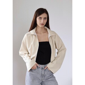 Op de afbeelding: Cr&egrave;mekleurige jas met kraag en ritssluiting, gecombineerd met een zwarte top en lichtblauwe jeans. De jas heeft lange mouwen en een relaxte pasvorm. Het model draagt de jas.
