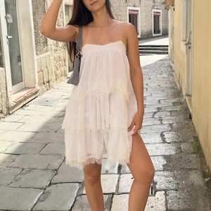 Pode incluir: Um vestido branco sem mangas com camadas, al&ccedil;as finas e um delicado acabamento em renda nas bordas de cada n&iacute;vel. O vestido apresenta uma silhueta solta e fluida com um decote reto e &eacute; feito de um tecido macio e leve.