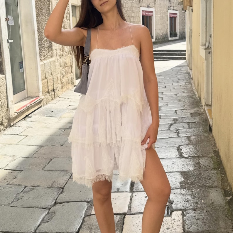 Pode incluir: Um vestido branco sem mangas com camadas, al&ccedil;as finas e um delicado acabamento em renda nas bordas de cada n&iacute;vel. O vestido apresenta uma silhueta solta e fluida com um decote reto e &eacute; feito de um tecido macio e leve.