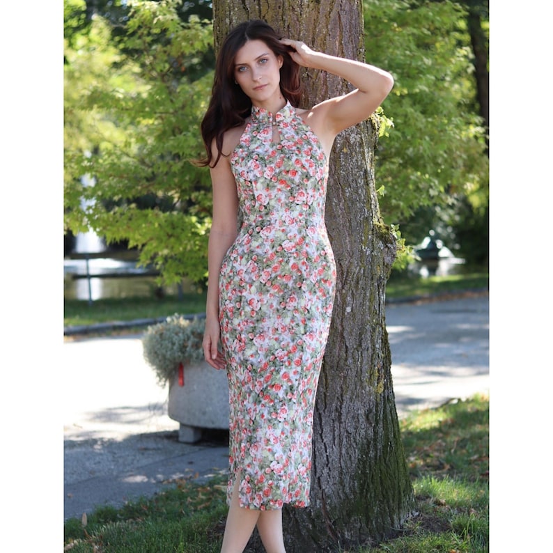 K&ouml;nnte beinhalten: &Auml;rmelloses Kleid mit Blumenmuster und hohem Ausschnitt. Das Kleid hat ein Muster aus roten und rosa Blumen mit gr&uuml;nen Bl&auml;ttern auf wei&szlig;em Grund. Das Kleid ist knielang und figurbetont.