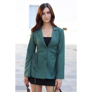 May include: A green blazer with a cinched waist and long sleeves. The blazer has a button closure and tie details on the sides. The model is wearing a black top and a black skirt.