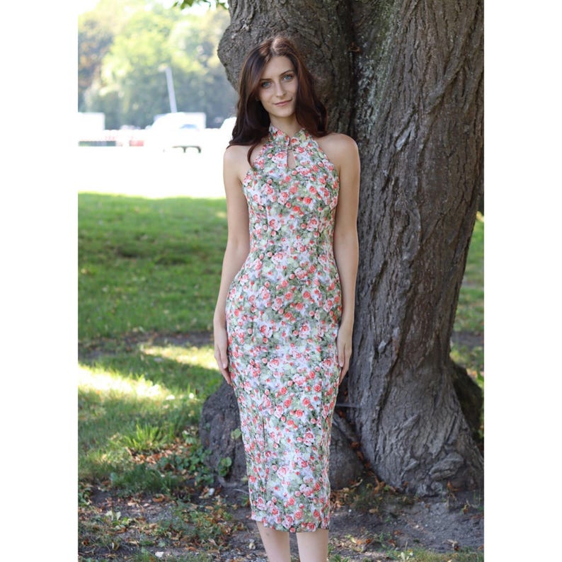 K&ouml;nnte beinhalten: Ein halterneck-Kleid mit Blumendruck und Schl&uuml;sselloch-Ausschnitt. Das Kleid hat ein Muster aus roten und rosa Rosen auf hellem Hintergrund. Das Kleid ist knielang und figurbetont.