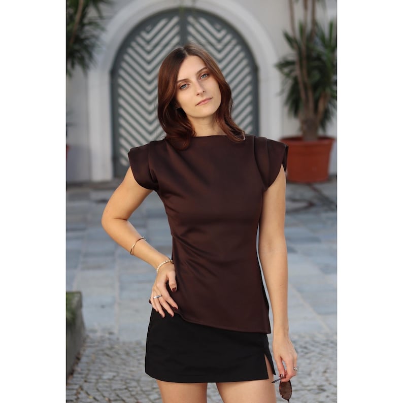 May include: A brown, short-sleeved top with a boat neck and a black skirt. The top has a fitted silhouette and structured shoulders. The skirt is a mini-skirt with a side slit. The outfit is worn by a person.