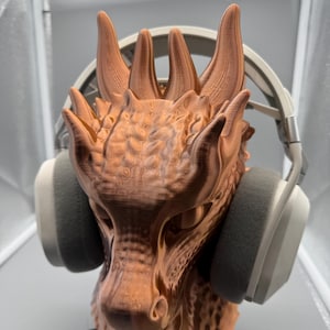 May include: A copper-coloured dragon head sculpture with a headset resting on it. The dragon has detailed scales and horns. The headset has grey earcups and a white headband. A black cord extends from the base.