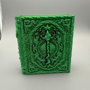 May include: A green, elaborately decorated book with detailed relief carvings. The cover features a central design with a staff and serpent, surrounded by floral and geometric patterns. The book's edges are also intricately detailed.