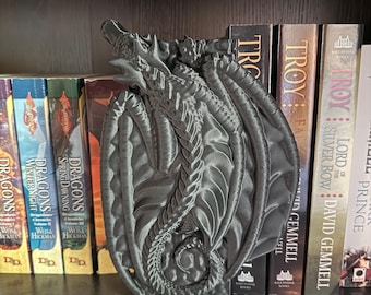 May include: A gray dragon-shaped bookend, with detailed wings and scales, stands on a shelf next to a row of books. The dragon is positioned to hold books upright, adding a fantasy element to the bookshelf.