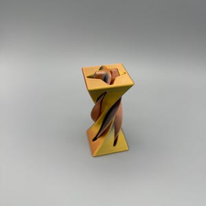 Puede incluir: Una escultura abstracta decorativa con una parte superior e inferior cuadradas, y un diseño geométrico retorcido. La escultura presenta un degradado de colores, que transita del dorado al cobre y al marrón. La obra de arte se exhibe sobre un fondo gris neutro.
