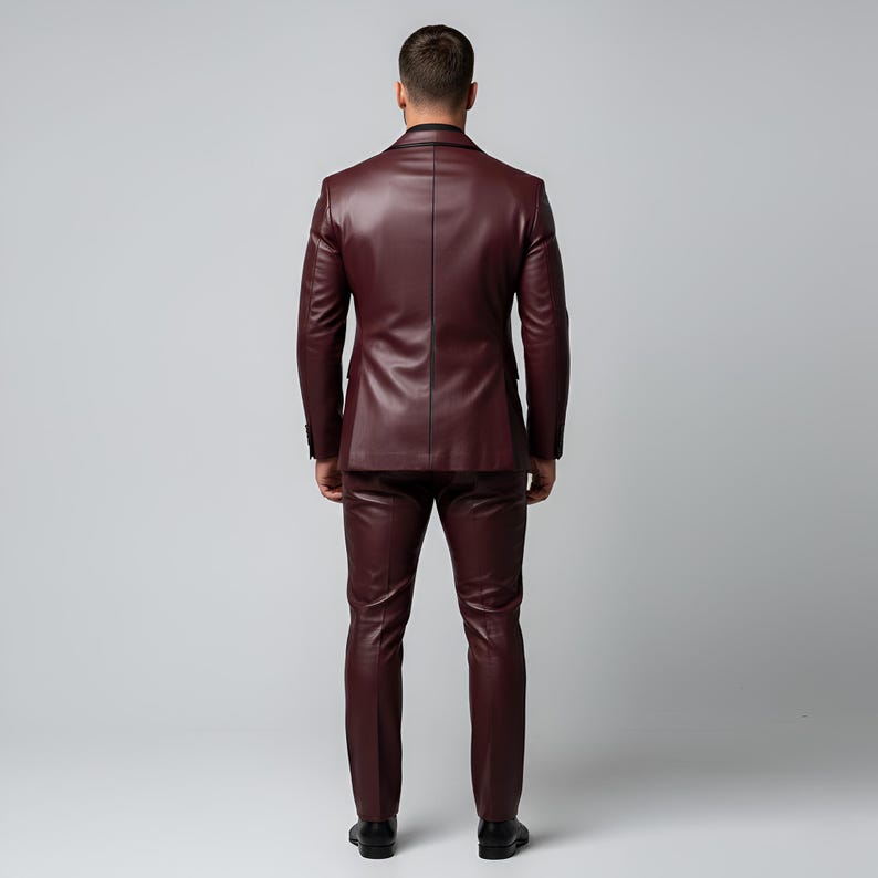 K&ouml;nnte beinhalten: Ein burgunderfarbener Lederanzug, bestehend aus einer ma&szlig;geschneiderten Jacke und einer passenden Hose. Die Jacke hat einen klassischen Zwei-Knopf-Verschluss und fallende Revers. Die Hose ist schmal geschnitten. Der Anzug wird mit einem schwarzen Hemd und schwarzen Schuhen getragen.