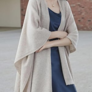 May include: A beige shawl-style cardigan with wide sleeves. The cardigan is draped over a dark blue dress. The outfit is neutral-toned and suitable for layering. The cardigan has a simple, elegant design.