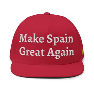 May include: A red snapback cap featuring the white embroidered text Make Spain Great Again on the front panel. The cap includes a small Spanish flag patch on the side, a structured crown, and a flat brim, providing a classic headwear design.